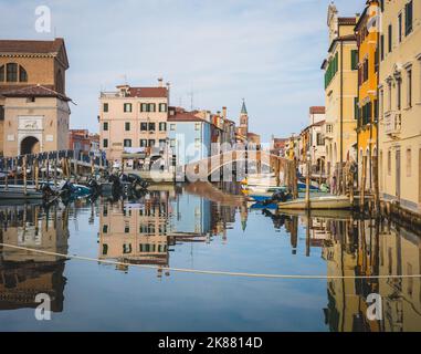 Chioggia cityscape with narrow water canal with moored boats, buildings, brick bridge and tower of San Giacomo Apostolo church – Venetian lagoon,Italy Stock Photo