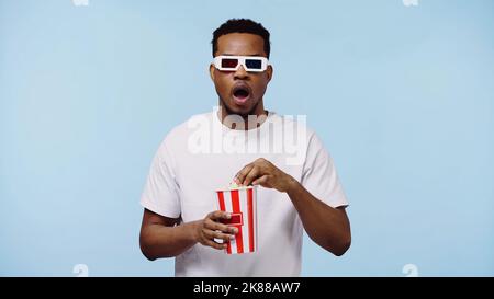 surprised african american man in 3d glasses watching movie and holding popcorn bucket isolated on blue Stock Photo
