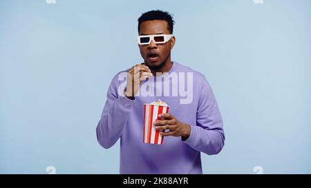 shocked african american man in 3d glasses watching movie and holding popcorn bucket isolated on blue Stock Photo