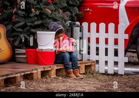 Little African American boy in bear costume sits on wooden pallet near the Christmas tree, opens gift on the street. In the backyard There is a red re Stock Photo