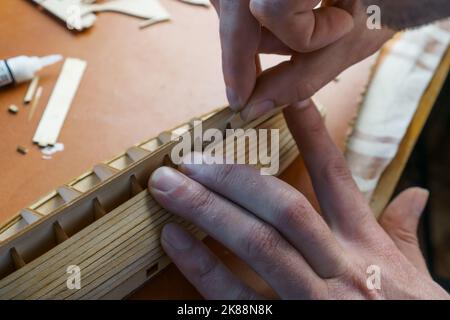 Hands of man gluing plywood details for ship model with glue, holding with fingers. Process of building toy ship, hobby, handicraft. Table with Stock Photo