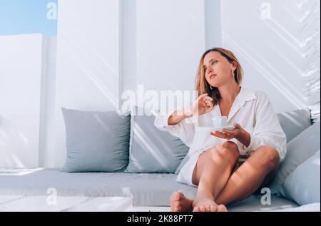 Beautiful barefoot woman dressed white shirt relaxing on luxury terrace with espresso cup patio on the comfortable sofa pillows and enjoying midday ti Stock Photo