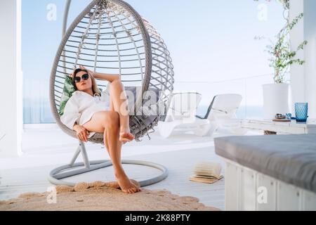 Beautiful woman in fancy black sunglasses lying in a comfortable hanging chair on the open house terrace and enjoying lazy middle day hours. Modern te Stock Photo