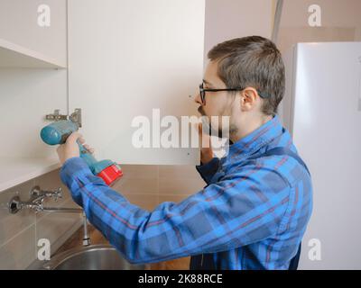 young man in blue work suit using electric screwdriver when assembling cupboard in kitchen. Home renovation concept. Stock Photo