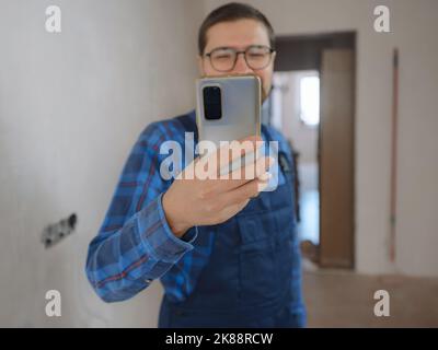 young man in blue work suit doing Repair apartment. Home renovation concept. Guy with smartphone texting and tapping on cellphone, searching for solut Stock Photo