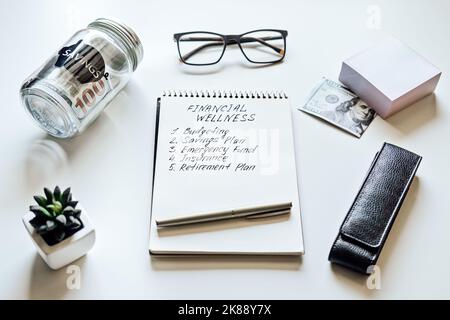 Financial Wellness. Open notepad with text Financial Wellness and list on table with mason jar saving bank, stationery and glasses. Investment in life Stock Photo