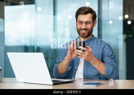 Successful and happy businessman with beard works in modern bright office, male boss uses laptop, business owner in casual shirt holds phone, browses internet pages Stock Photo