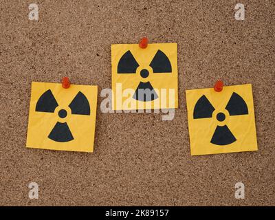 Three yellow paper notes with radiation warning signs on them pinned to a corkboard. Close up. Stock Photo