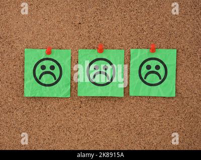 Three green paper notes with sad faces on them pinned to a corkboard. Close up. Stock Photo
