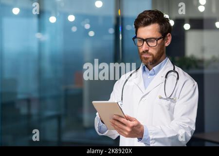 A serious and thoughtful doctor reads medical information from a tablet computer, a man in a medical gown uses a tablet, concentrates, types messages and reviews the patient's record Stock Photo