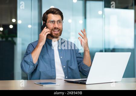 Successful freelancer working inside modern office building, man talking on phone, businessman using laptop at work Stock Photo