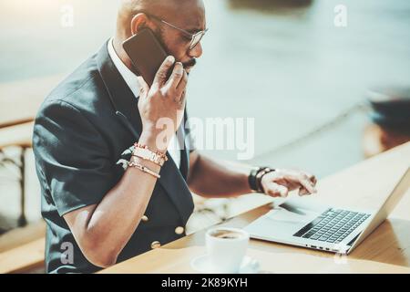 A side view of a serious black businessman talking on the phone while sitting with his netbook in a street cafe; an African man freelancer on a patio Stock Photo