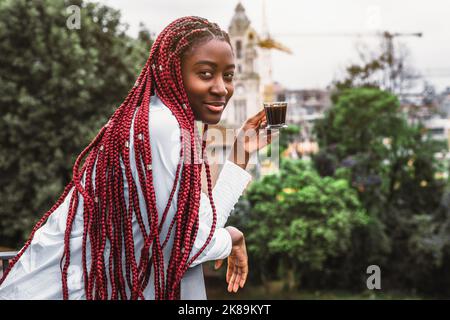 A portrait of a ravishing youthful black female with long red braids enjoying her morning espresso coffee while leaning against the fencing of a balco Stock Photo