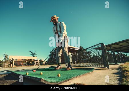 View of a dapper African guy with a black beard and a cigar in his mouth, in a straw hat, he is ready to hit the ball using a club during a golf train Stock Photo