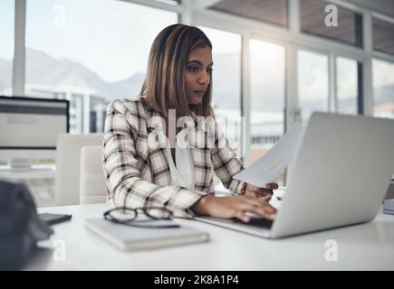 Multitasking has always been a strong suit. an attractive young businesswoman sitting alone in her office and using her laptop while reading paperwork Stock Photo