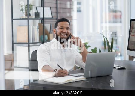 I never keep success waiting on the line. Portrait of a young businessman talking on a cellphone while writing notes and working on a laptop. Stock Photo