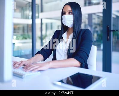 Get everything in place to keep you safe. a young businesswoman wearing a face mask and using a computer in a modern office. Stock Photo
