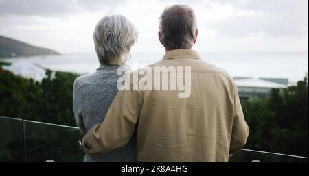 Love turns a grey day into a great day. Rearview shot of a happy senior couple standing together on a balcony and looking at the ocean view. Stock Photo