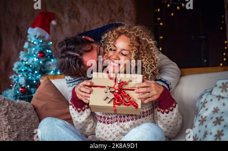 Happiness in christmas holiday eve season. Man doing a surprise to happy woman giving her a xmas gift in a red decorated box. Christmas tree in backgr Stock Photo
