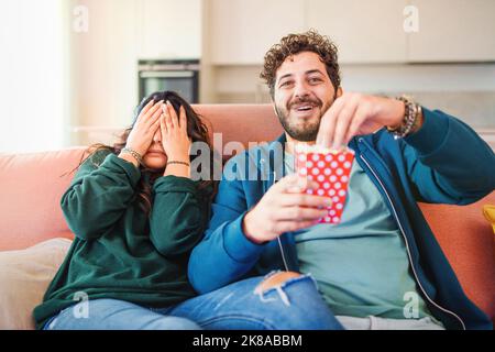 Scared millennial newlywed couple watching horror movie on tv, frightened young woman covering eyes and man laughing and eating popcorn during a thril Stock Photo