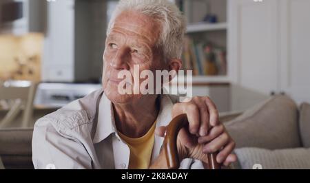Every day is the same. a senior man sitting alone on the sofa at home and looking contemplative while holding his walking stick. Stock Photo