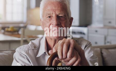 My walking stick is my retirement buddy. a senior man sitting alone on the sofa at home and holding his walking stick. Stock Photo