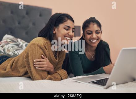 What do you want to watch. two young women using a laptop while lying together on a bed. Stock Photo