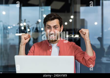 Close up of successful businessman, man celebrating victory and successful triumph, looking at laptop screen reading good news and happy holding hands up, winner gesture Stock Photo
