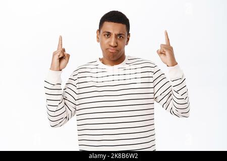 Annoyed african american man points up, sulks and looks bothered by smth, stands over white background Stock Photo