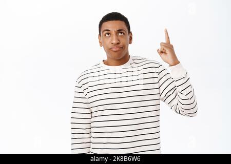 Annoyed african american man points up, sulks and looks bothered by smth, stands over white background Stock Photo