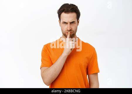Angry man shows shh, shush sign, press finger to lips with frowned scary face, keep quiet gesture, stands over white background Stock Photo