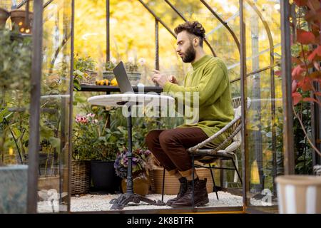 Man works remotely on laptop in glasshouse at backyard Stock Photo