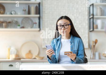 Portrait of a young beautiful Asian woman holding a credit card and a phone in her hand. Sitting at home in the kitchen, looking at the camera. Stock Photo