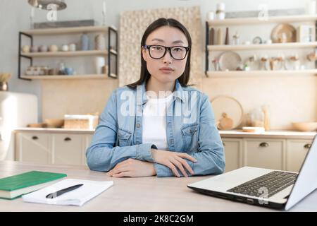 Portrait of a young Asian businesswoman working at home and conducting online interviews from a laptop. A serious, purposeful woman is sitting at the table, looking at the camera. Stock Photo