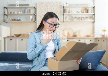 Happy and pleasantly surprised young beautiful Asian woman sitting on sofa at home and opening cardboard box, gift. I received an unexpected package in the mail. Stock Photo