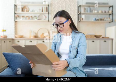 A young beautiful Asian woman received a package by a courier with home delivery. Sitting on the sofa at home and opening the box, online order from the store. Stock Photo