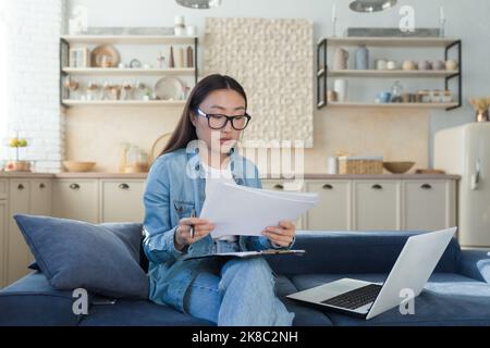Young beautiful creative woman Asian writer, journalist. He sits at home on the sofa with a laptop and writes a book, an essay, a script, an article. Holds paper and pen. Stock Photo