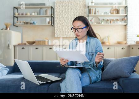 Work at home online. Busy young Asian woman architect, designer works at home. She sits focused on the sofa with a laptop, projects, and a pen. Stock Photo