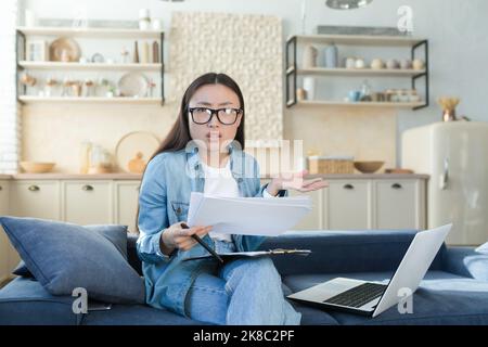 Confused and nervous young Asian woman sitting on sofa at home and working with documents and laptop. Checks the accounts and finances of the family budget. He looks at the camera, spreads his hands. Stock Photo