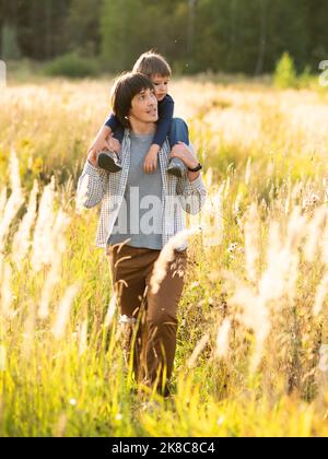 Father carries his son on shoulders. Family time outdoors. Man with child on autumn field at sunset. Dad and son explores nature together. Stock Photo