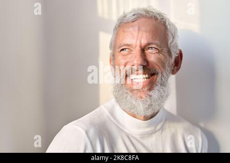 Smiling bearded senior old man laughing standing at white wall in sunlight. Stock Photo