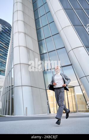 Happy stylish older business man wears suit running outdoor in big city street. Stock Photo