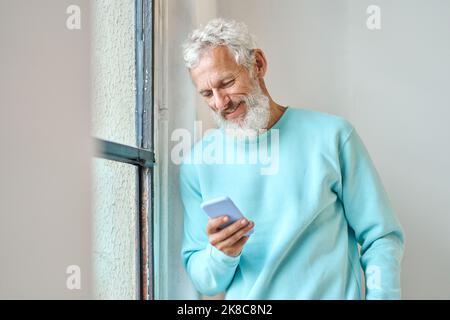 Smiling old bearded hipster man using mobile phone standing at window. Stock Photo