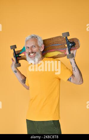 Happy bearded senior man skater holding skateboard isolated on yellow. Stock Photo