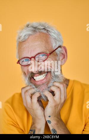 Happy funny older hipster man wearing glasses touching beard isolated on yellow. Stock Photo
