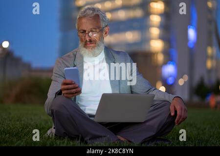 Old business man sitting outside office in city park using smartphone at night. Stock Photo
