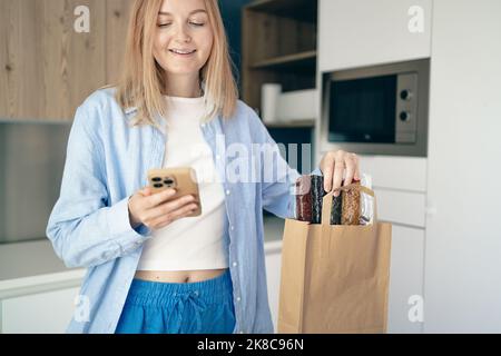 Young woman unpacking paper bag after shopping using smartphone in kitchen interior at home, copy space. Stock Photo