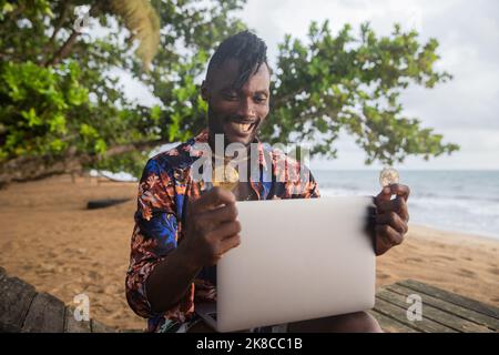 An African cryptocurrency trader checks the market with his laptop on the beach. Stock Photo