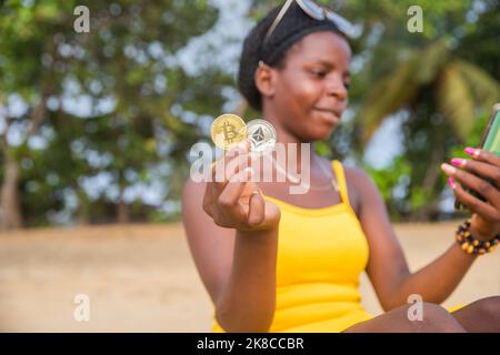 An african trader with Bitcoin and ethereum coins on the beach checks the market with her phone Stock Photo