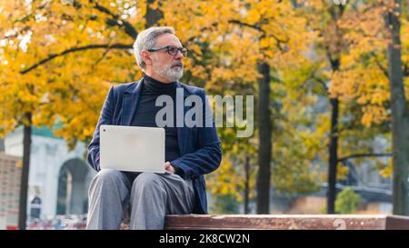 Modern stylish bearded grey-haired mature man entrepreneur sitting in the park with fall foliage and working on his laptop. High quality photo Stock Photo